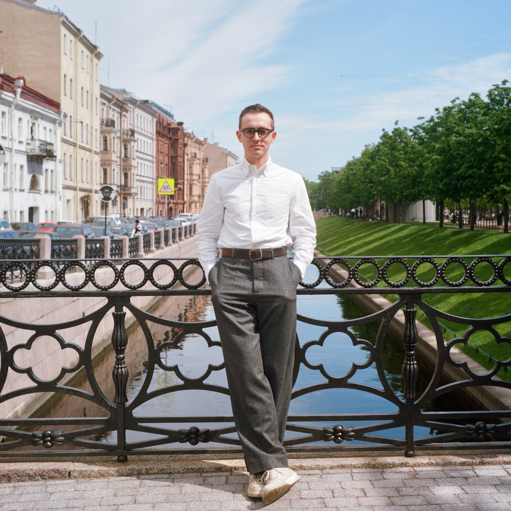 Full size photograph of a young man wearing glasses, white shirt, grey trousers and white sneakers, standing on a bridge across the canal